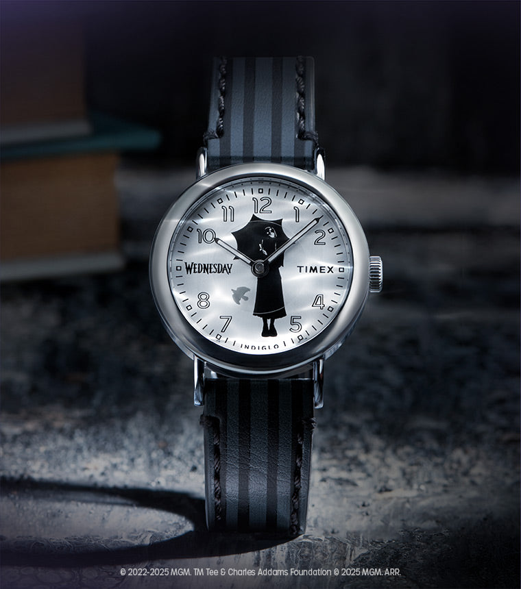 Flatlay of the Timex x Wednesday watch resting on a dark surface with a blurred book in the background, evoking a mysterious, academic aesthetic.