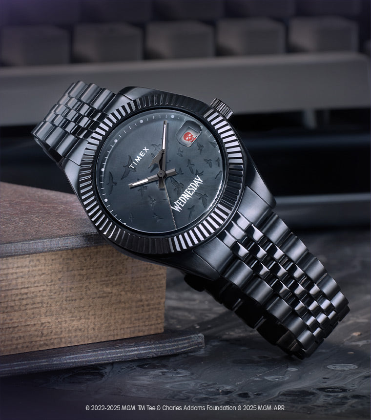 The Timex x Wednesday Legacy watch resting atop a stack of old books, with a vintage typewriter in the background. The watch features a black stainless-steel case and bracelet, a dark textured dial with scattered raven silhouettes, a skull icon at the date window, and “Wednesday” printed at the 6 o’clock position.