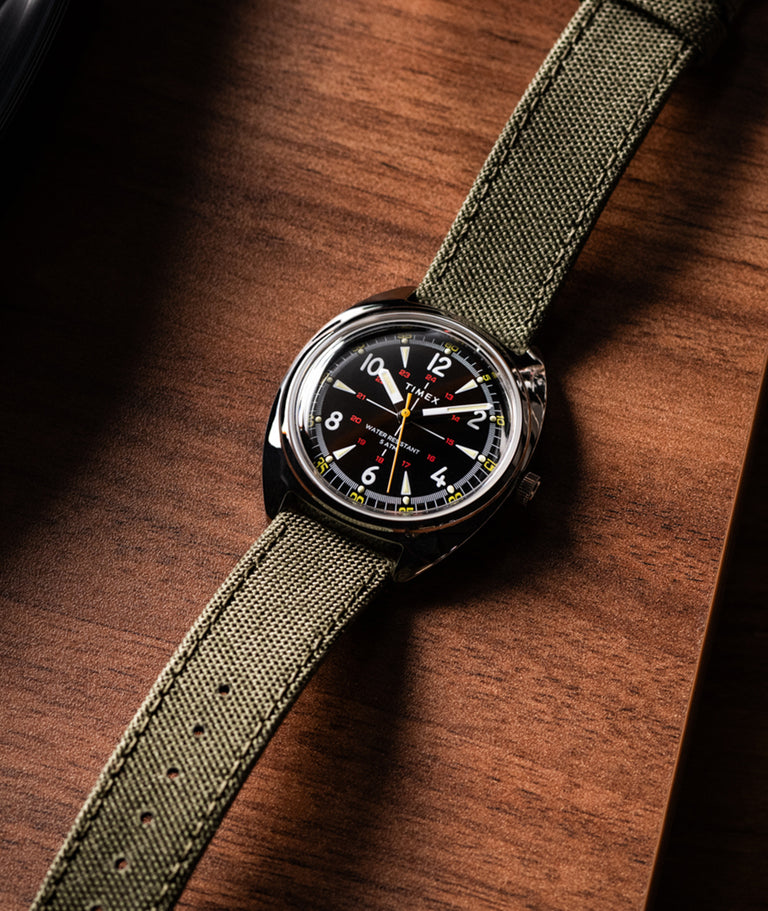Timex x Bespoke Post limited-edition watch resting on a wooden surface next to a vinyl record player. The timepiece features a bold black dial with large white numerals, red accents, and a yellow seconds hand. Housed in a polished cushion-shaped case, it is paired with an olive green fabric strap that adds a rugged, military-inspired touch. The overall aesthetic merges vintage design with modern functionality.