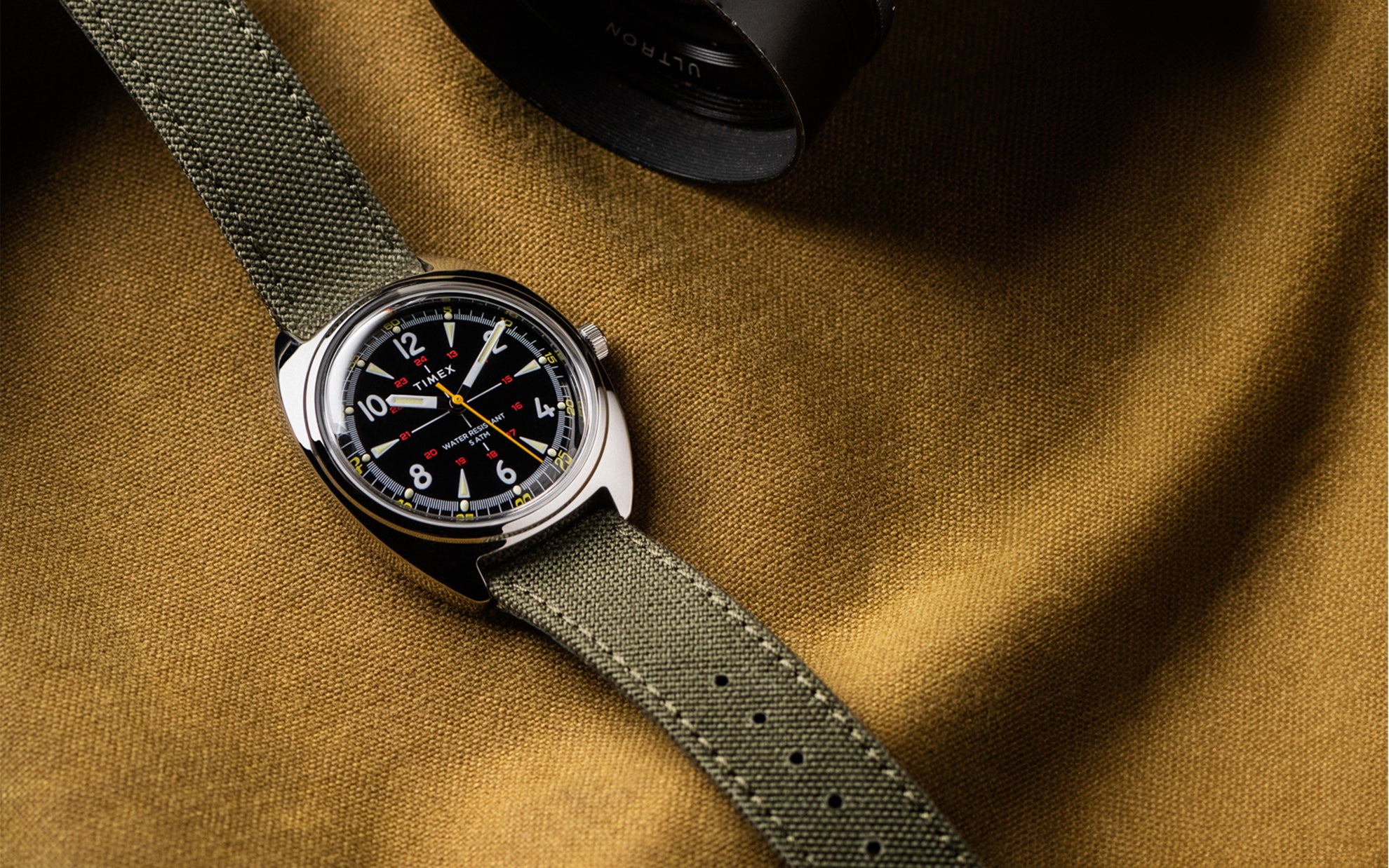 Timex x Bespoke Post watch featuring a black dial, yellow seconds hand, and olive green fabric strap, displayed flat on a brown textured fabric surface with a camera lens partially visible in the frame.