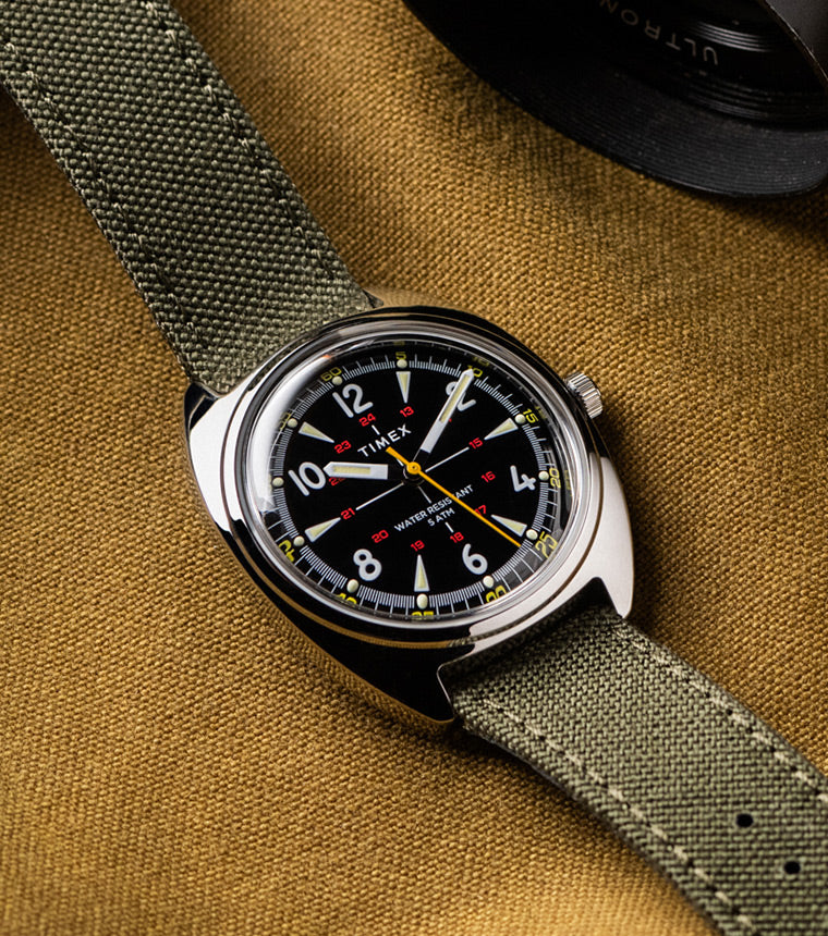 Timex x Bespoke Post watch featuring a black dial, yellow seconds hand, and olive green fabric strap, displayed flat on a brown textured fabric surface with a camera lens partially visible in the frame.