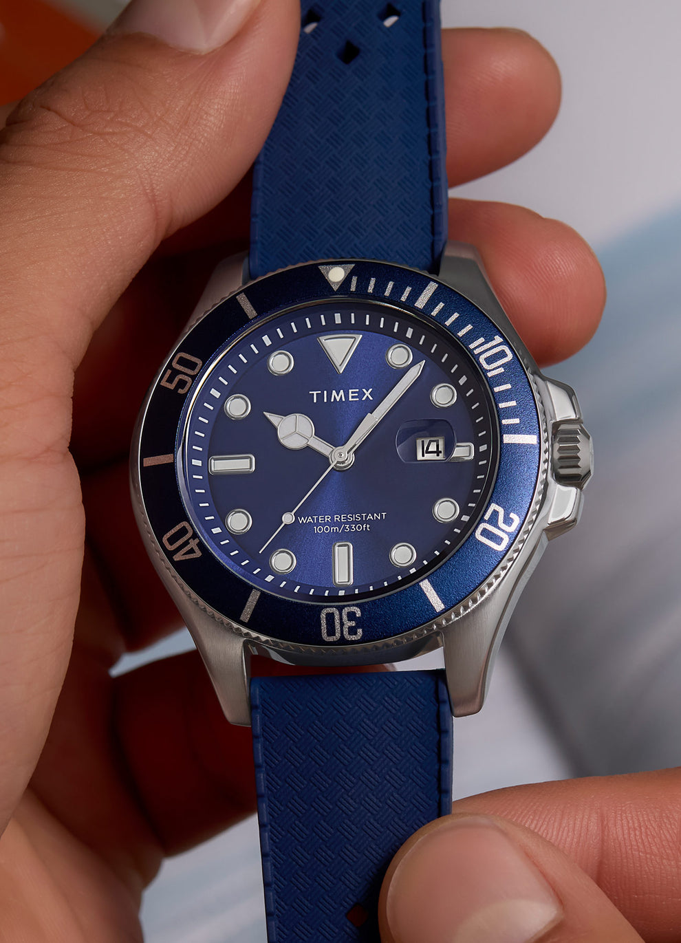 Close-up of a hand holding the Timex Harborside Coast watch, revealing the vibrant blue sunray dial and unidirectional bezel. The magnified date display at 3 o'clock and textured strap are clearly visible, highlighting the durable, ocean-inspired design.