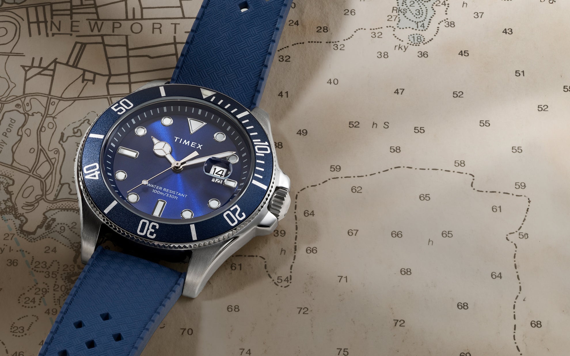 A Timex Harborside Coast watch with a navy blue silicone strap rests on a nautical chart map, showing the coast near Newport. The stainless steel case and blue dial reflect soft light, and the watch face reads “Water Resistant 100m / 330ft” with a magnified date window at 3 o'clock.