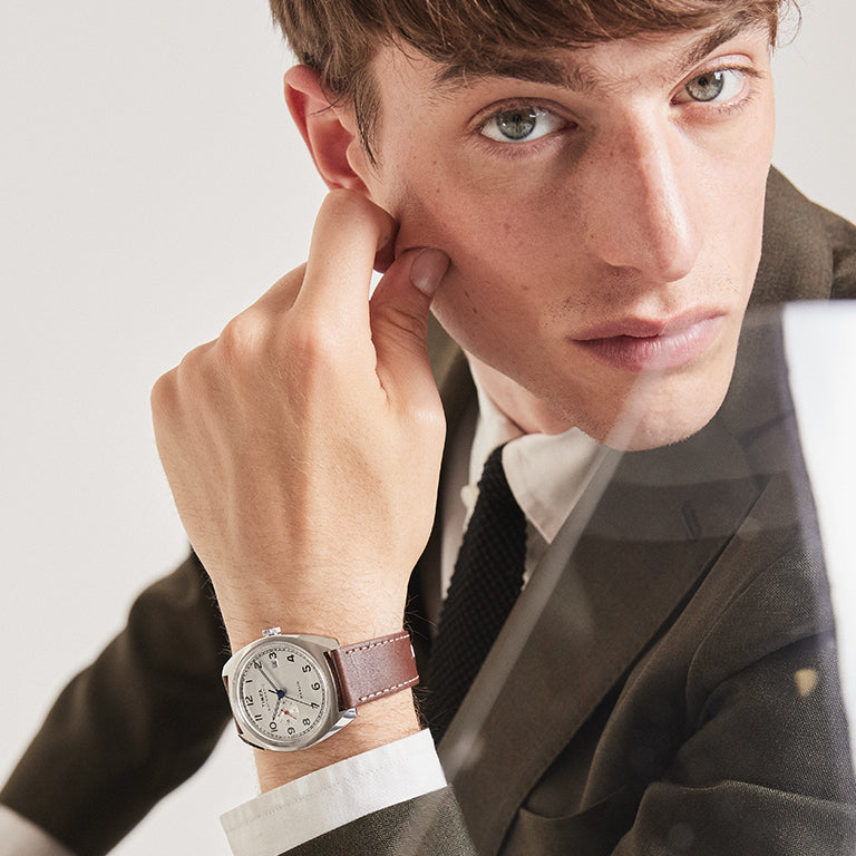 A young man dressed in a dark olive suit, white shirt, and black tie poses with his face resting against his hand. He is wearing a stylish wristwatch with a round silver case, white dial, and brown leather strap. The watch features a clean analog display with black numerals and a blue second hand.