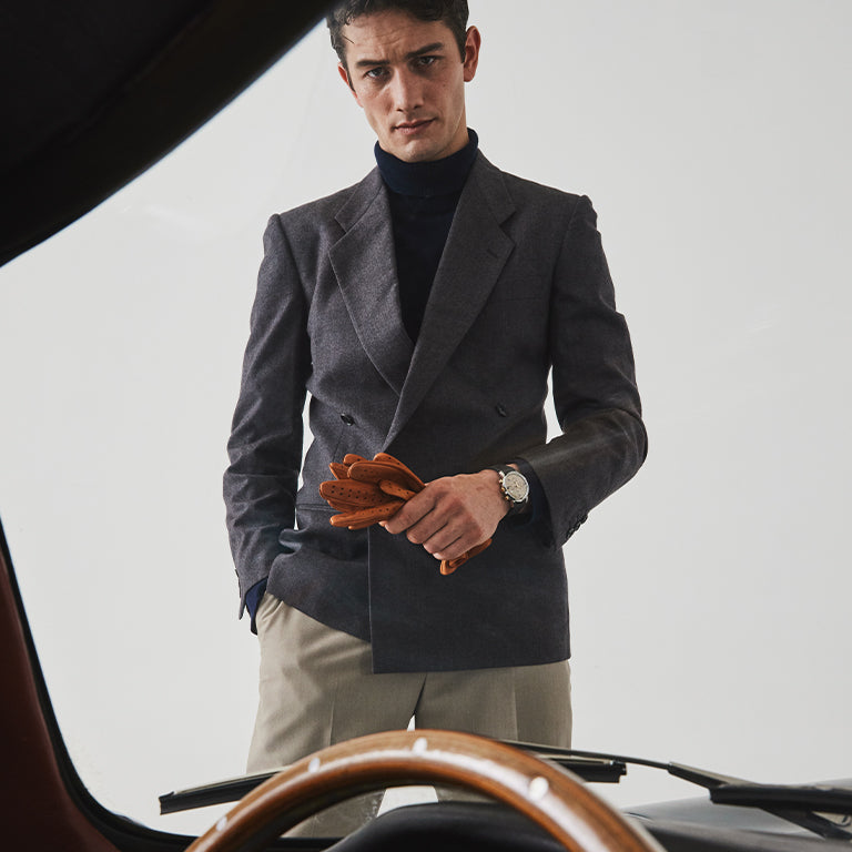 A stylish man stands in front of a vintage car, viewed partially from behind the steering wheel. He wears a tailored charcoal double-breasted blazer over a navy turtleneck, paired with beige trousers. In his hands, he holds a pair of tan leather driving gloves.
