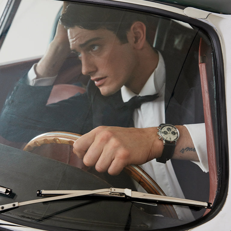 A man in a tuxedo sits inside a classic car, gripping the wooden steering wheel with one hand and leaning his head on the other in a thoughtful pose. Seen through the windshield, he wears a black leather strap chronograph watch with a cream dial and dark subdials.