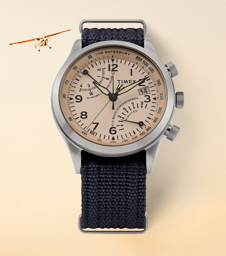 Lifestyle image of the Waterbury Traditional Fly-back Chronograph watch placed on a soft beige gradient background with a small vintage aircraft flying in the top left corner. The watch features a salmon-toned dial, bold black numerals, three sub-dials, and a navy-blue woven strap. The stainless steel case and aviation-inspired design evoke a pilot’s timepiece aesthetic.