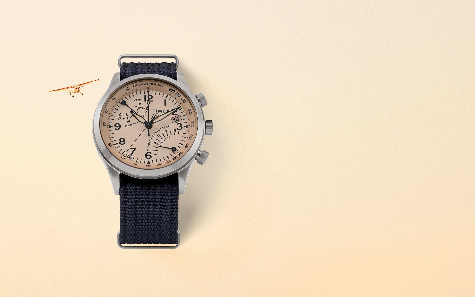 Lifestyle image of the Waterbury Traditional Fly-back Chronograph watch placed on a soft beige gradient background with a small vintage aircraft flying in the top left corner. The watch features a salmon-toned dial, bold black numerals, three sub-dials, and a navy-blue woven strap. The stainless steel case and aviation-inspired design evoke a pilot’s timepiece aesthetic.