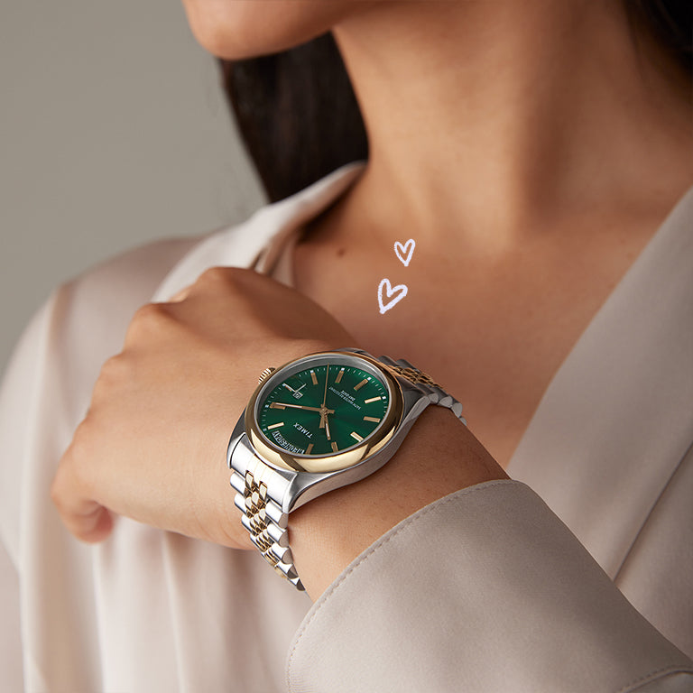 A close-up of a person wearing a classic Timex watch with a deep green sunray dial and a two-tone gold and silver bracelet. The person is dressed in a soft beige blouse, and two hand-drawn white hearts are drawn near the watch for a charming effect.