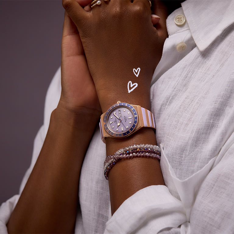 A close-up of a person wearing a stylish two-tone Timex watch with a lavender dial and diamond accents, paired with beaded bracelets. The person is dressed in a white textured blouse. Two hand-drawn white heart doodles float near the wrist, adding a playful touch.