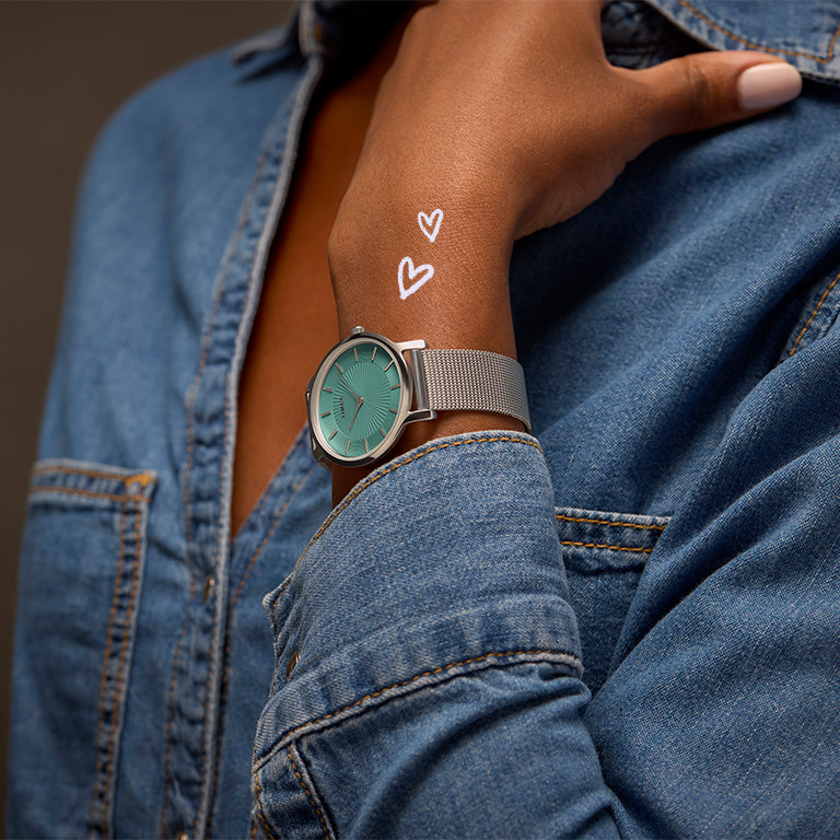 A close-up of a person wearing a sleek Timex watch with a teal dial and a silver mesh strap. The person is dressed in a denim jacket, and white hand-drawn hearts are illustrated near the wrist, giving a playful and stylish vibe.
