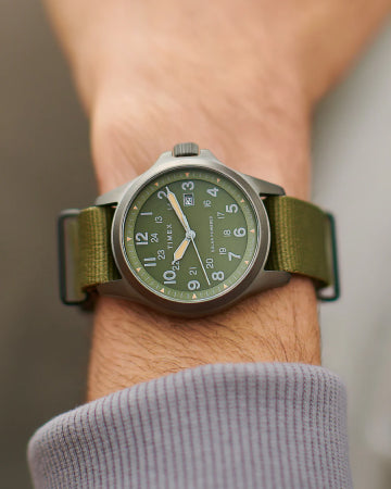 A close-up of a Timex solar-powered wristwatch with a green dial and an olive green fabric strap. The watch features a stainless steel case, luminous hands, and a date display, blending a rugged and functional design suited for outdoor adventures. The watch is worn over a light gray long-sleeve shirt, emphasizing its casual yet durable style.