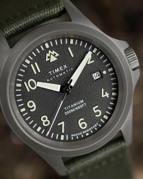 A close-up of a Timex Automatic Titanium wristwatch with a textured black dial, white numerals, and luminous hands. The watch features a date display, a durable titanium case, and a rugged olive green fabric strap. Designed for adventure, it is water-resistant up to 200 meters and shock-resistant, emphasizing durability and functionality.