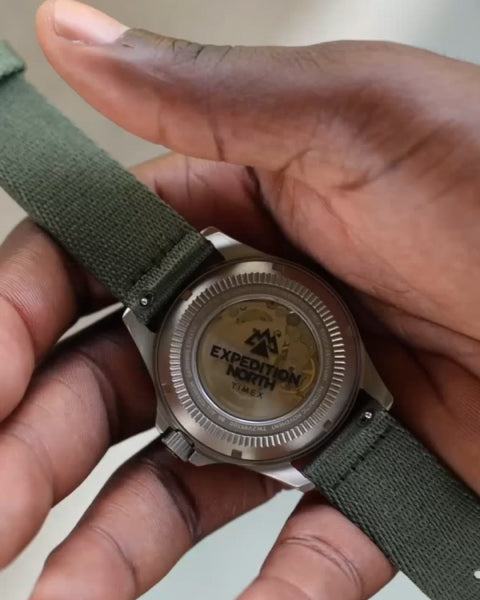 A close-up of a Timex Expedition North wristwatch held in a person's hand, showcasing the transparent case back. The exposed mechanical movement is visible through the clear back, with 'Expedition North' branding etched on the glass. The watch features a titanium case and an olive green fabric strap, emphasizing durability and an adventure-ready design.