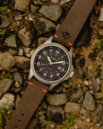 A Timex expedition-style wristwatch with a black dial, white numerals, and a date display, laid on a rugged gravel surface. The watch features a stainless steel case and a brown textured strap with orange stitching and an embossed logo, emphasizing its durability and outdoor-ready design.