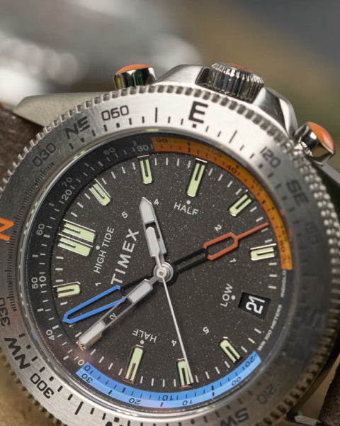A close-up shot of a Timex expedition-style wristwatch with a black speckled dial, luminescent markers, and multiple indicators, including a tide tracker. The stainless steel bezel features directional markings, and the watch has an adventurous, rugged design.