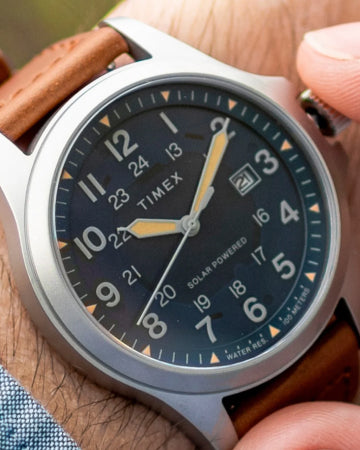 A close-up of a Timex solar-powered wristwatch with a black dial, luminous hands, and a date display. The watch features a stainless steel case and a brown leather strap, combining durability with a classic outdoor aesthetic. The wearer is adjusting the watch, highlighting its sleek and functional design.