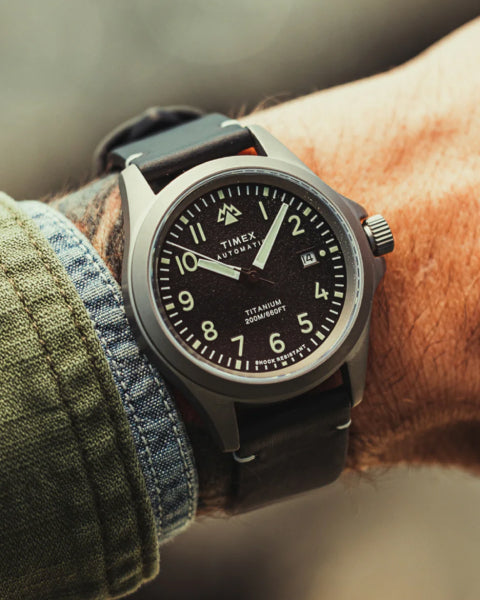 A close-up of a Timex Titanium Automatic wristwatch on a person's wrist. The watch features a black dial with white numerals, a date display, and a durable titanium case. It is paired with a black leather strap, complementing the rugged and adventure-ready aesthetic. The wearer is dressed in a green and denim jacket, adding to the outdoorsy style.