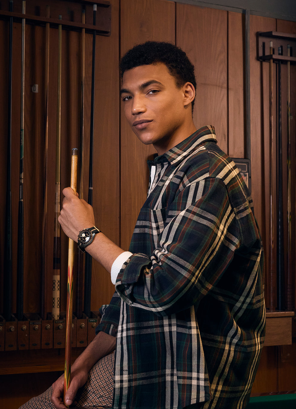 Lifestyle image of a male model in a plaid shirt holding a pool cue while wearing the Timex Waterbury Heritage Chronograph watch, set in a retro pool hall environment with wooden wall paneling and cue racks.