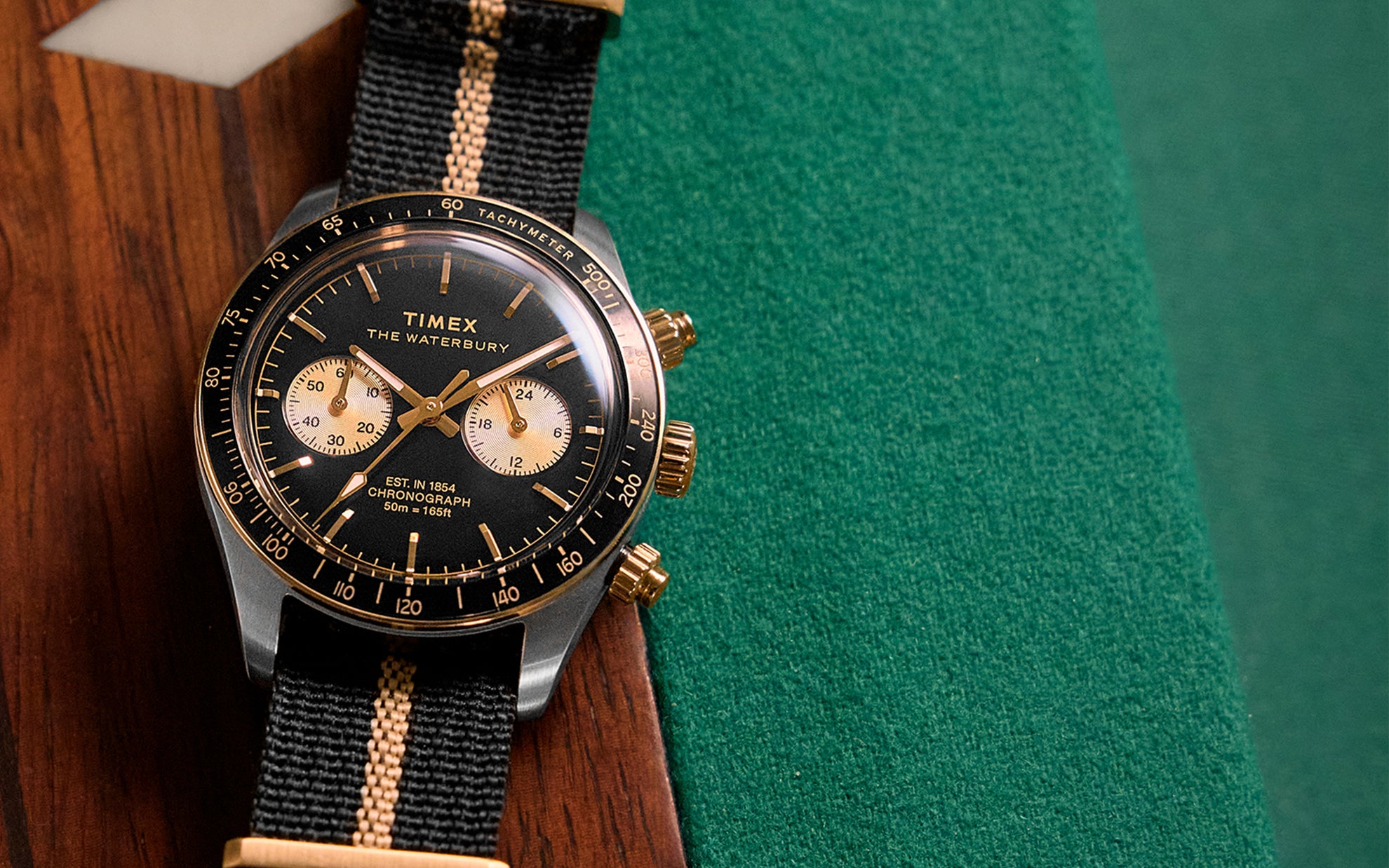 Close-up lifestyle image of the Timex Waterbury Heritage Chronograph wristwatch, featuring a black dial with gold accents, two sub-dials, and a black-and-tan striped fabric strap, placed stylishly on a wooden and green felt surface.