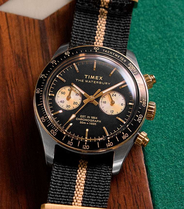 Close-up lifestyle image of the Timex Waterbury Heritage Chronograph wristwatch, featuring a black dial with gold accents, two sub-dials, and a black-and-tan striped fabric strap, placed stylishly on a wooden and green felt surface.