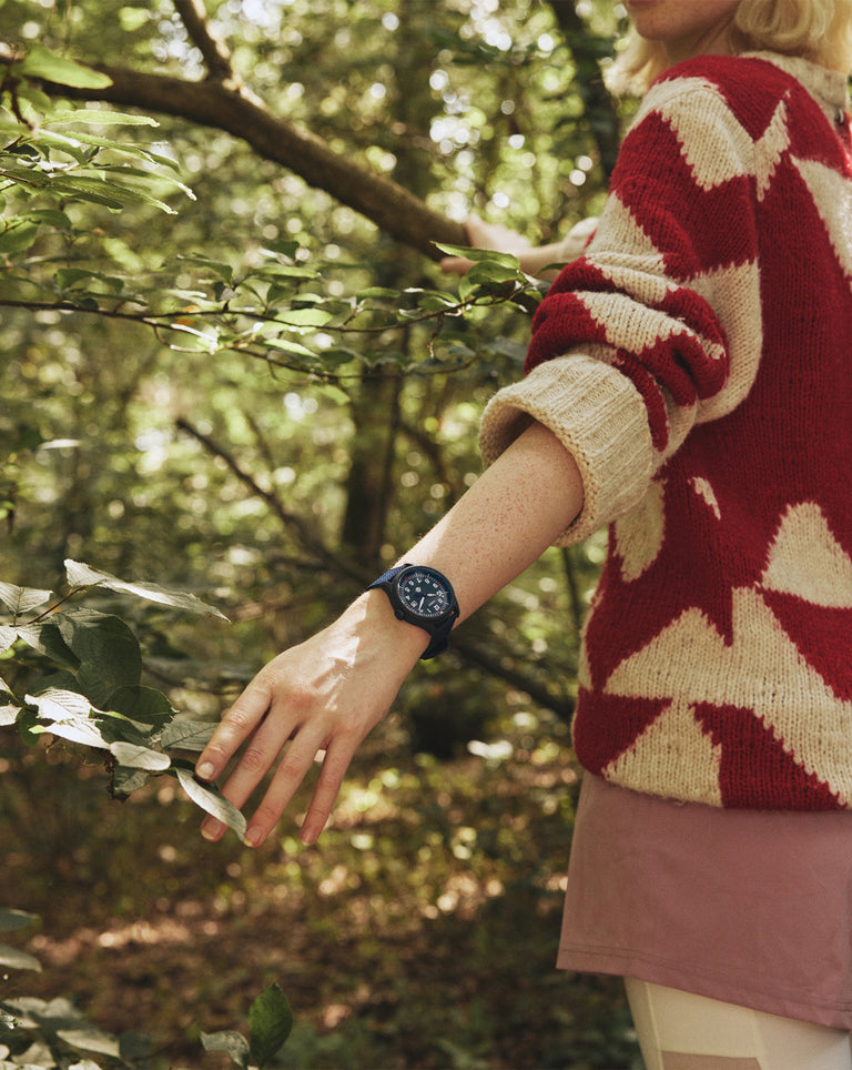 A woman explores a wooded trail, wearing a red and beige geometric-patterned sweater and a navy blue field watch. Her hand gently brushes past tree branches as she walks through the forest.