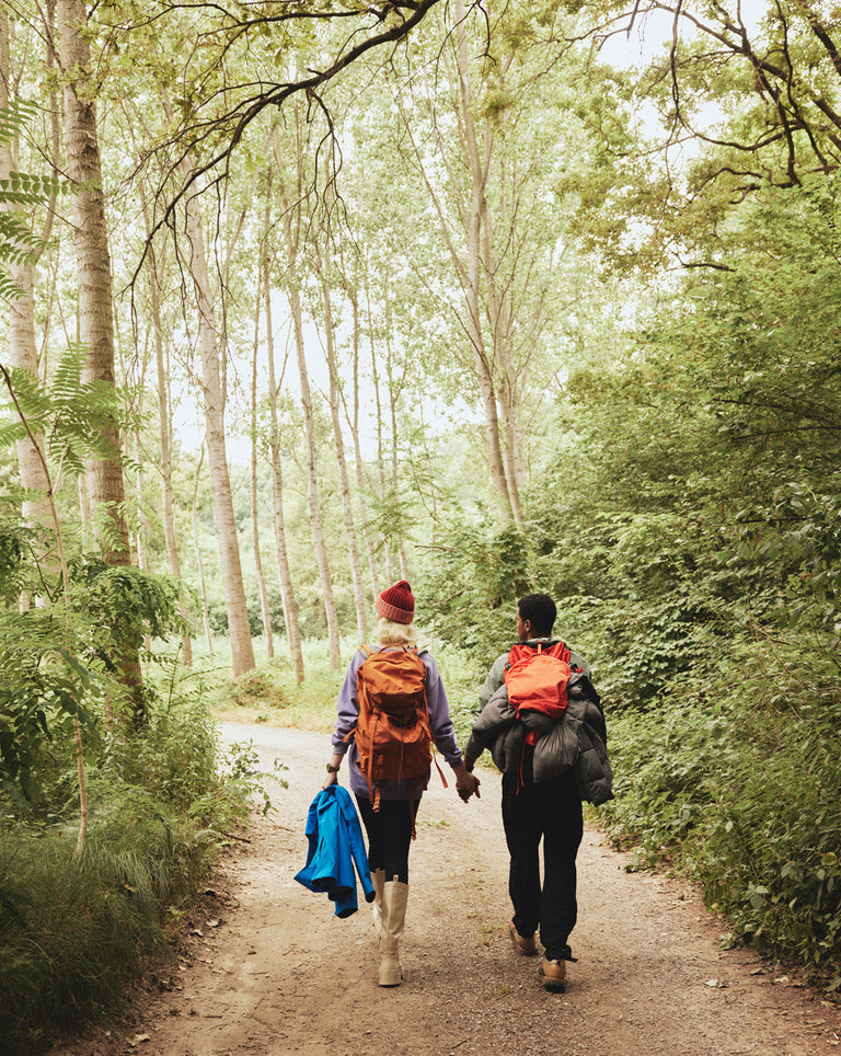 Two hikers, a man and a woman, walk hand-in-hand along a wooded trail surrounded by lush green trees. Both carry large backpacks with outdoor gear; the woman wears light boots, a red beanie, and holds a blue jacket, while the man is dressed in dark clothing with a bright orange pack attached to his bag.