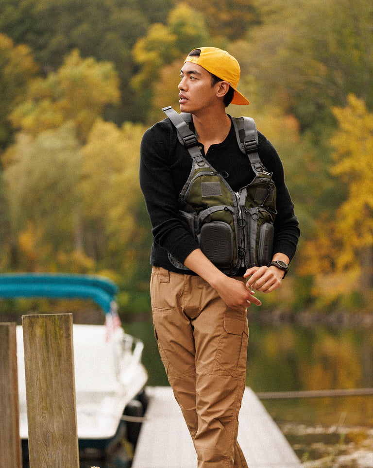 A young man stands on a dock beside a lake, dressed in tan cargo pants, a black long-sleeve shirt, and a bright yellow baseball cap worn backward. He sports a tactical fishing vest with gear pouches and wears a rugged wristwatch. Behind him, a white boat is docked, and the background is filled with vibrant autumn foliage, giving a warm and adventurous tone.