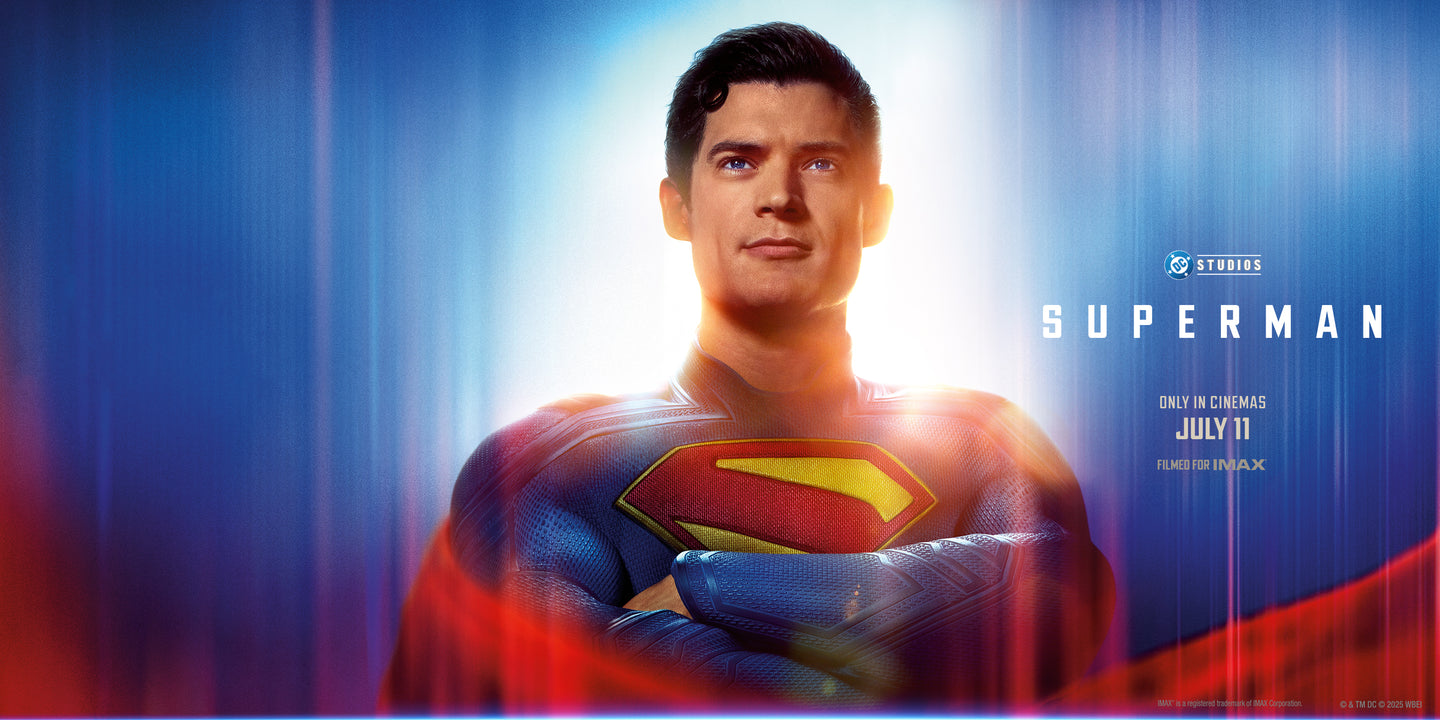 DC Studios’ Superman, featuring the superhero in a blue textured suit with the iconic red and yellow “S” emblem on his chest. He stands confidently with arms crossed, looking upward with a slight smile, illuminated by a bright light from behind. The background is a blend of blue and red streaks, with text reading “SUPERMAN” and “Only in cinemas July 11 – Filmed for IMAX.