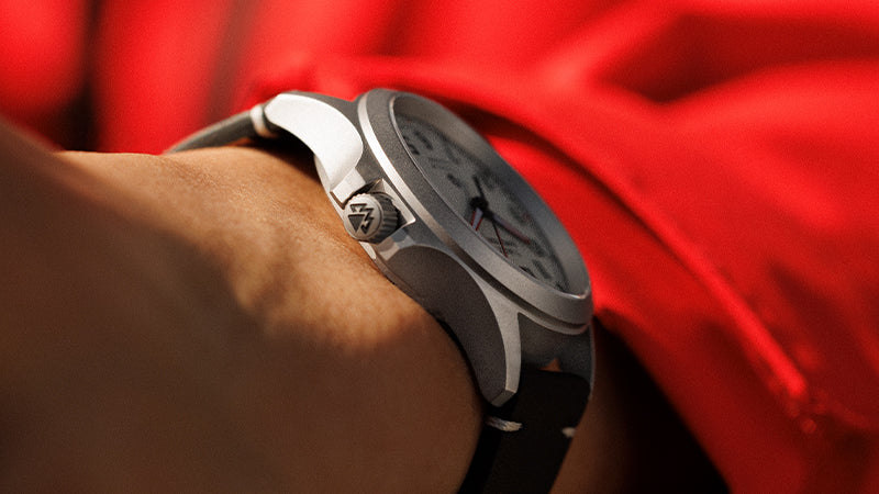 Side profile of a Timex Expedition watch showcasing the brushed titanium case and embossed crown logo, worn with a red sleeve visible in the background.