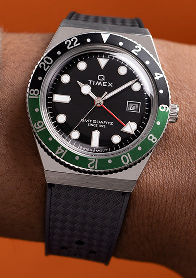 Close-up image of a Q Timex GMT wristwatch worn on the left wrist, featuring a black dial with bold white hour markers, a red GMT hand, and a date window at 3 o’clock. The bezel is split in black and green, marked with 24-hour numerals, emphasizing the dual time zone functionality. The watch is paired with a textured black strap and set against a vibrant orange background, highlighting the retro-sporty design.