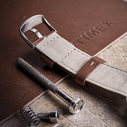 Close-up image of a beige and brown watch strap laid on a textured work surface alongside metal components, springs, and a leather mat embossed with the Timex logo. The scene emphasizes craftsmanship and material quality.