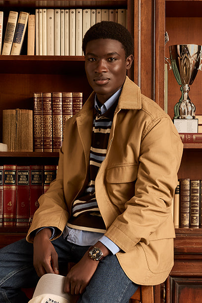 A well-dressed man sits confidently in front of a wooden bookshelf filled with books and a trophy. He wears a tan jacket over a striped shirt and displays a bold Timex chronograph watch on his wrist, blending heritage style with modern sophistication.