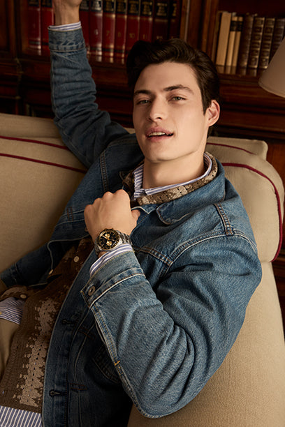 A man lounges comfortably on a beige couch in a library, smiling while showing off a Timex chronograph watch. He wears a denim jacket layered over a patterned sweater, embodying relaxed luxury and timeless style.