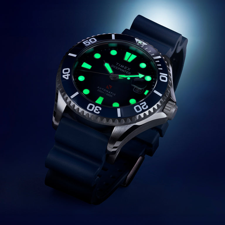 Timex Deepwater Automatic dive watch with luminous green markers and hands, unidirectional bezel, and navy silicone strap, glowing against a dark blue background.