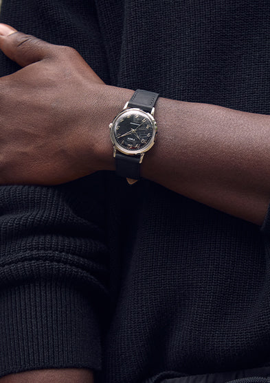 Close-up lifestyle image of a person wearing the Timex x Todd Snyder watch with a black dial and black leather strap, styled with a black textured sweater for a modern monochrome look.