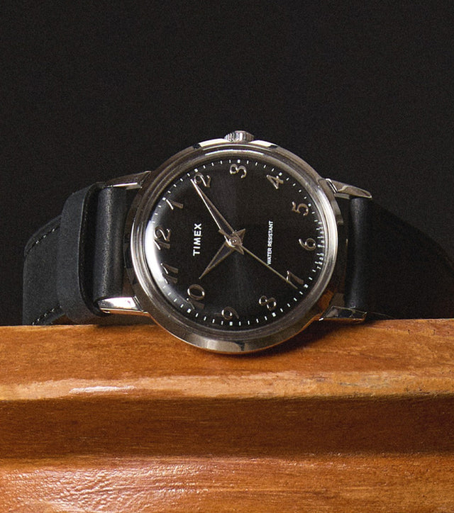 Timex x Todd Snyder watch resting on a wooden surface, highlighting the polished silver-tone case, black sunray dial, and classic Arabic numerals with a sleek black leather strap.