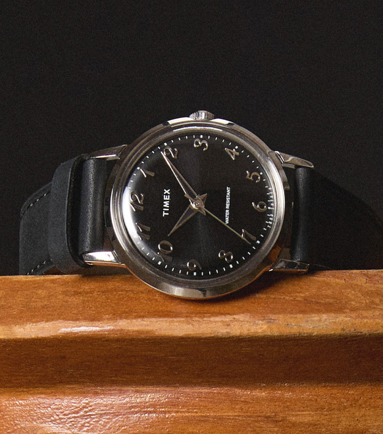 Timex x Todd Snyder watch resting on a wooden surface, highlighting the polished silver-tone case, black sunray dial, and classic Arabic numerals with a sleek black leather strap.