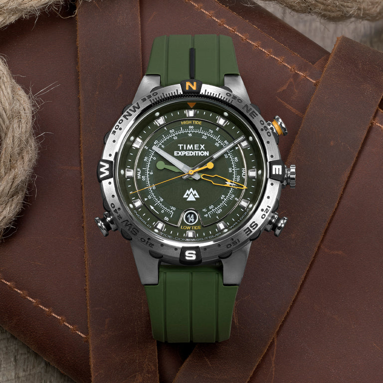 Timex Expedition watch with an olive green dial, tide-tracking features, compass bezel, and green silicone strap, displayed on rugged leather gear with rope accents.