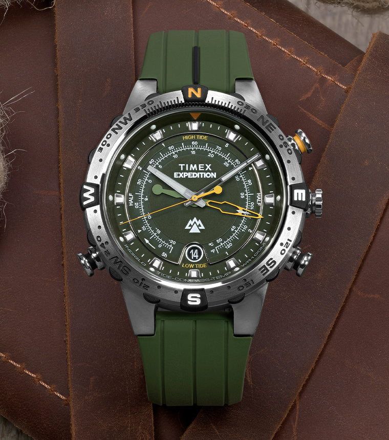 Timex Expedition® Field Post Solar watch with black dial, white numerals, date window at 3 o’clock, and black fabric strap, resting on a brown leather surface.