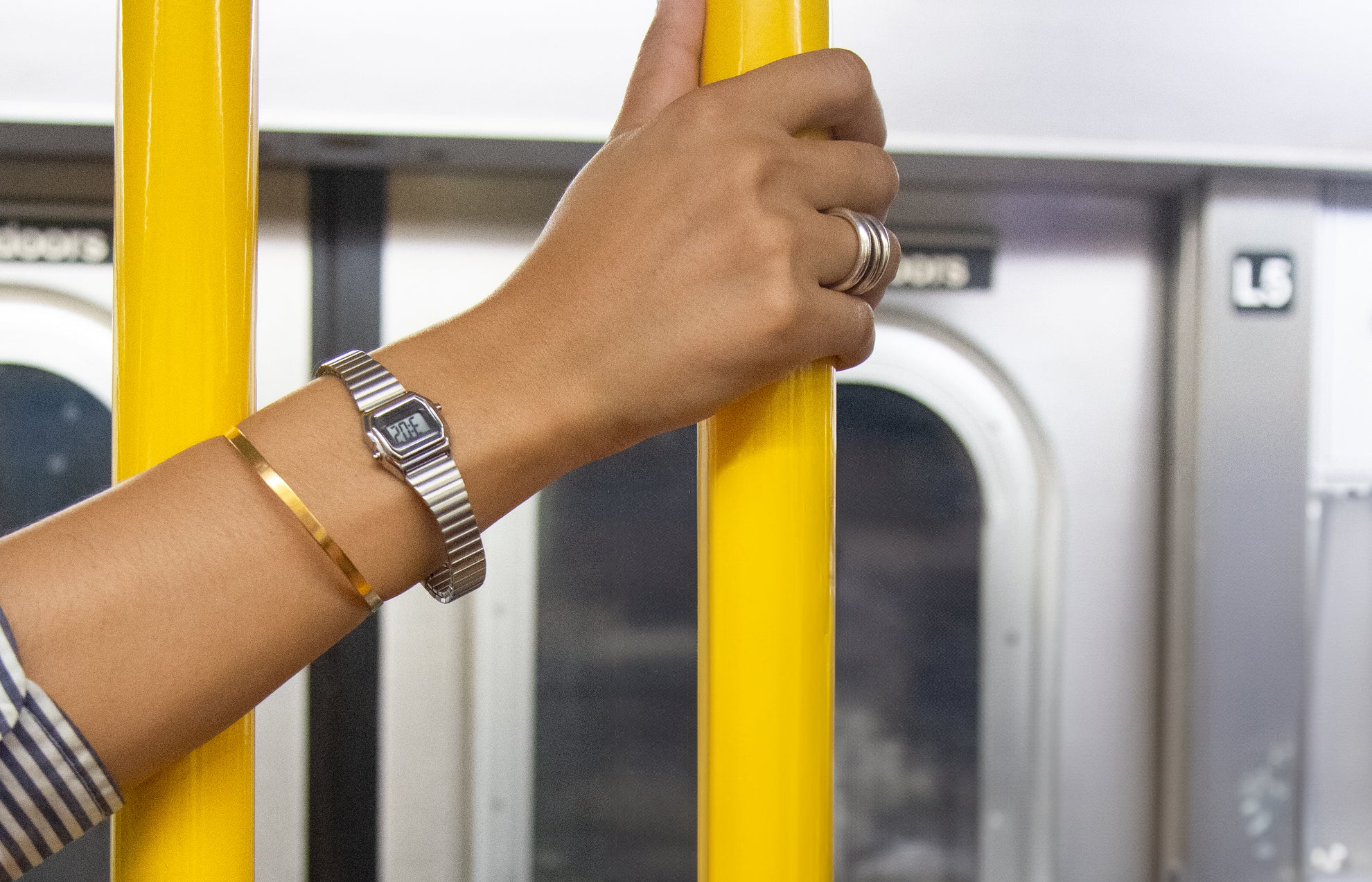 A person wearing the Teeny Tiny Timex digital watch with a silver-tone expansion band holds a yellow subway pole. The sleek, retro mini digital display complements a gold bangle, highlighting a minimalist city commute aesthetic.