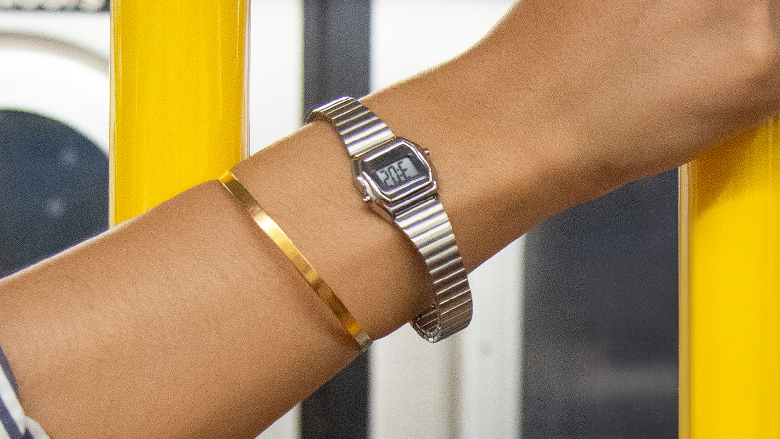 A person wearing the Teeny Tiny Timex digital watch with a silver-tone expansion band holds a yellow subway pole. The sleek, retro mini digital display complements a gold bangle, highlighting a minimalist city commute aesthetic.