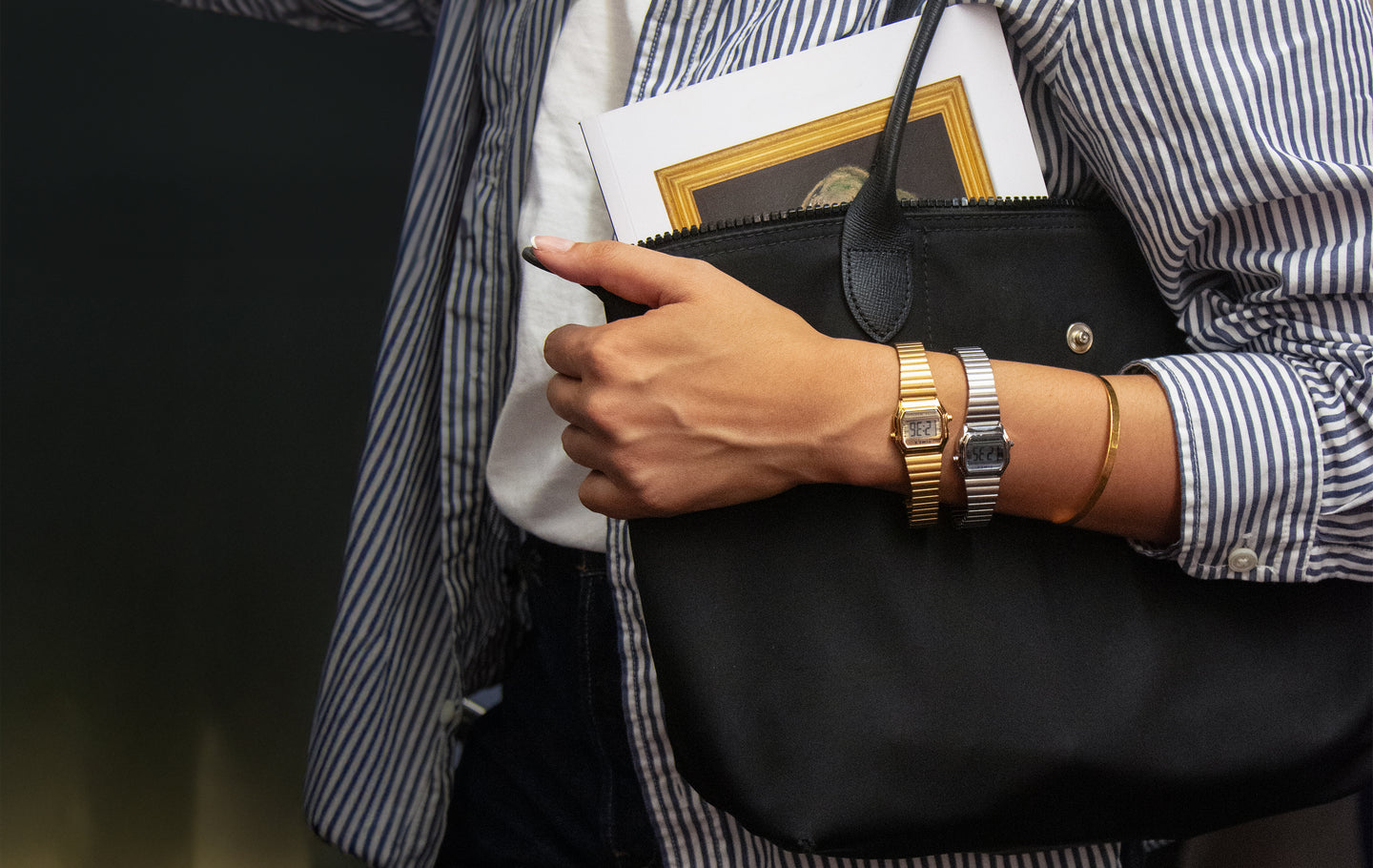 A person wearing two small rectangular Timex digital watches — one gold and one silver — on their wrist, along with a thin gold bracelet. They are holding a black handbag and a small framed print, dressed in a blue-and-white striped shirt over a white top.