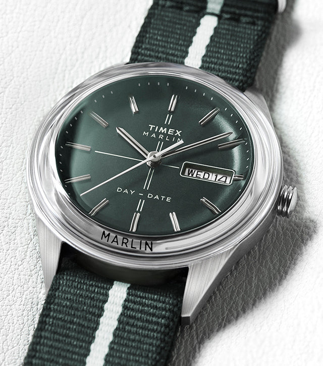 Timex Marlin® Jet Quartz watch with a deep green dial, silver-tone markers, and day-date window, paired with a dark green fabric strap featuring a white center stripe, displayed on a light leather surface.
