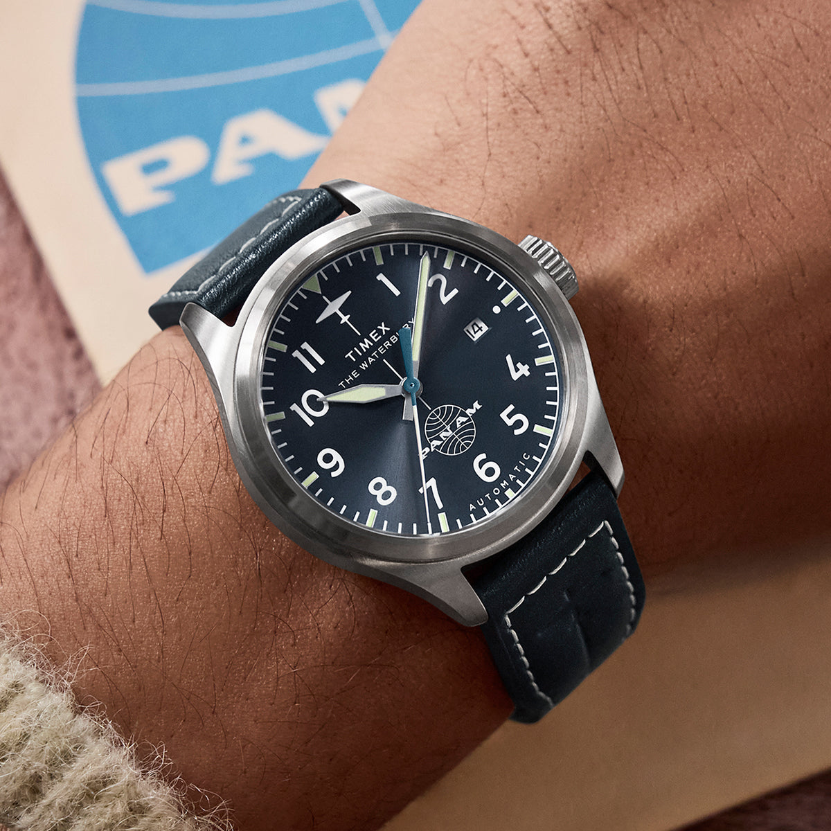 Timex x Pan Am® Waterbury Ace automatic watch on wrist, showcasing the navy dial, white Arabic numerals, date window, and Pan Am globe logo above 6 o’clock.