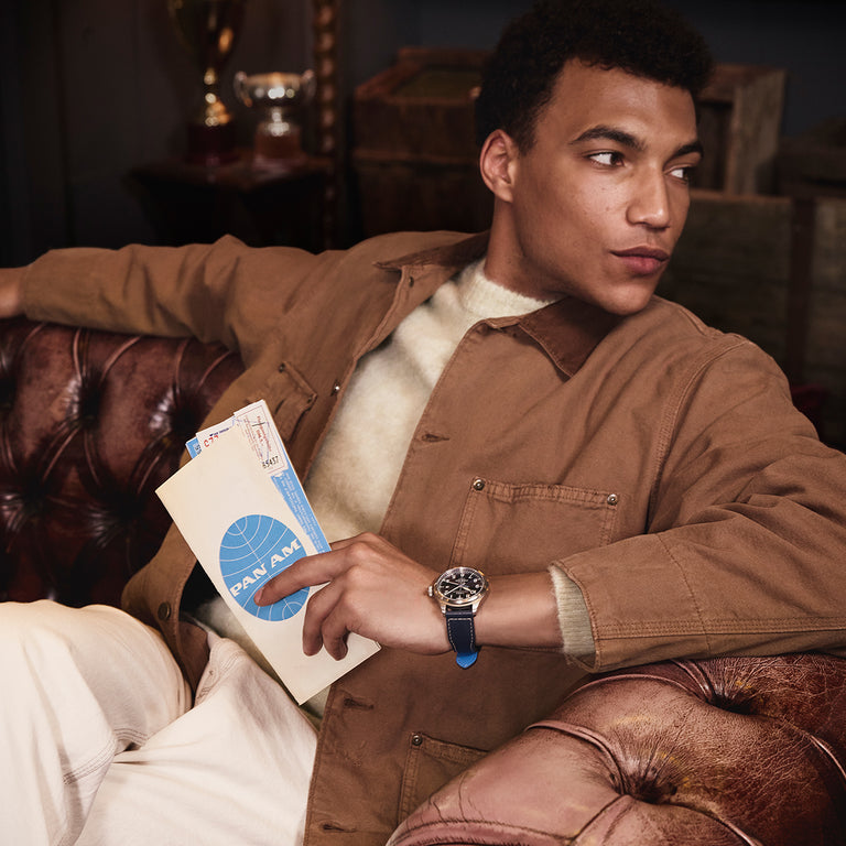 A man sitting on a brown leather couch holds a vintage Pan Am envelope and ticket in one hand while wearing a Timex x Pan Am watch with a blue strap. He’s dressed in a tan jacket and light sweater, looking off to the side in a warmly lit, retro-styled room.