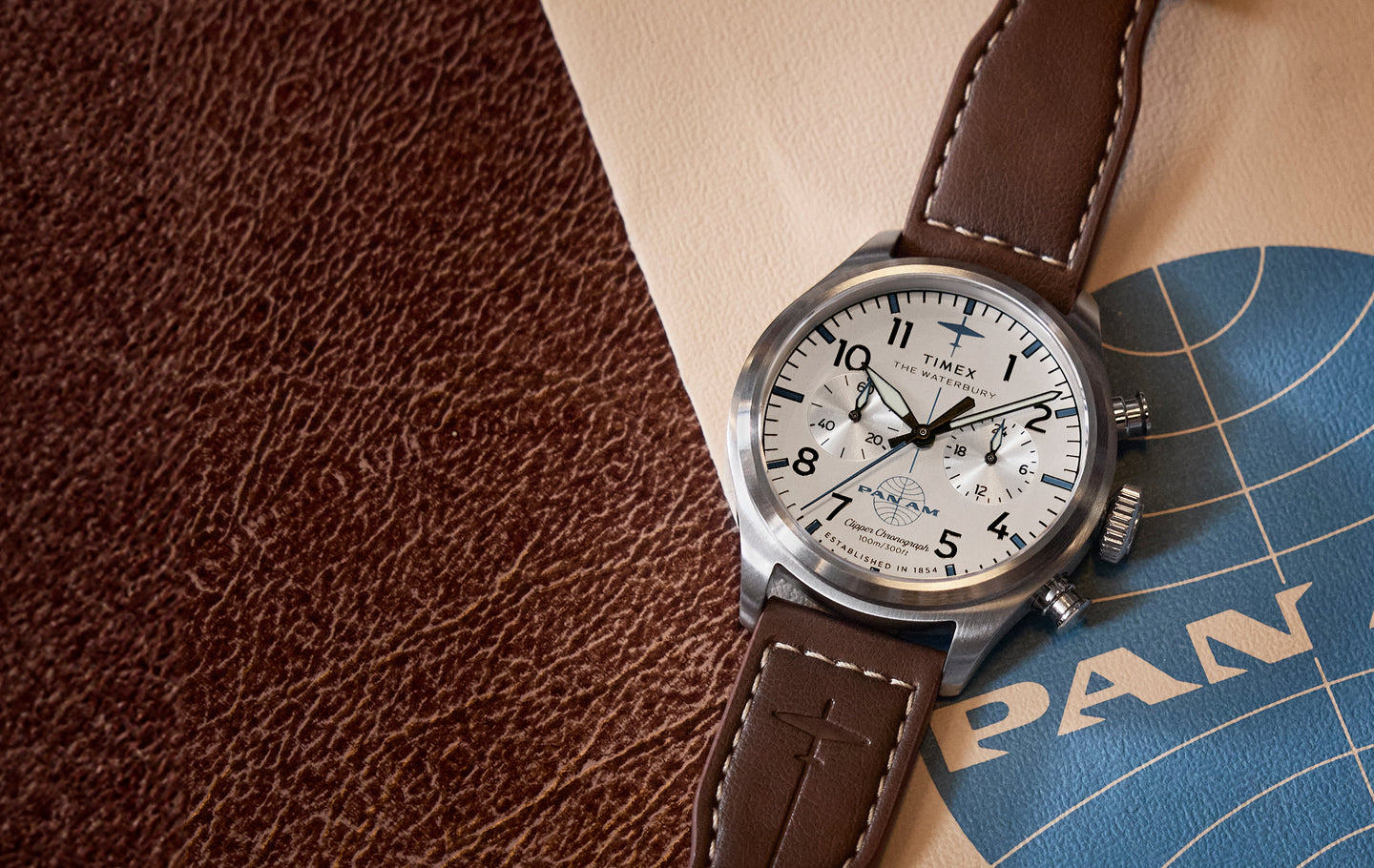 Timex x Pan Am® Waterbury Ace Chronograph watch with a white dial, black numerals, and brown leather strap, placed on vintage-style Pan Am branding and textured leather background.
