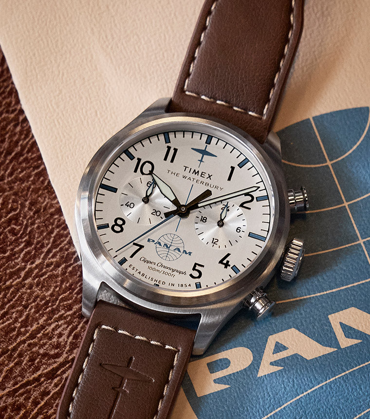 Timex x Pan Am® Waterbury Ace Chronograph watch with a white dial, black numerals, and brown leather strap, placed on vintage-style Pan Am branding and textured leather background.