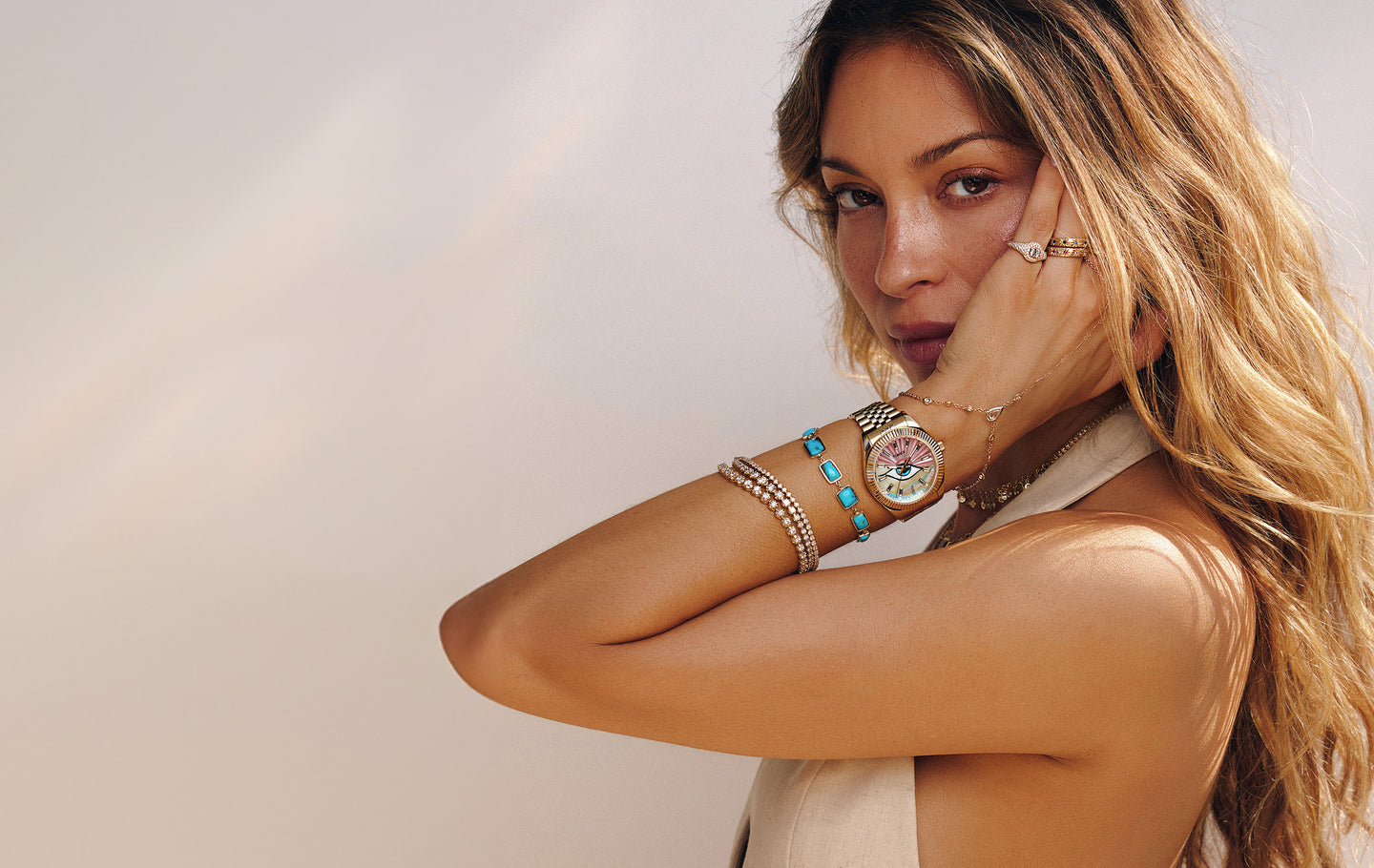 Woman wearing the Timex x Jacquie Aiche gold watch with a pink and multicolored mother-of-pearl dial featuring an eye motif, styled with layered gold and turquoise bracelets, posing with her hand near her face against a soft neutral background.