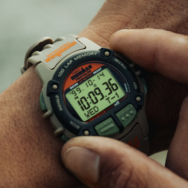 Close-up of the Timex x Huckberry Ironman Triathlon digital watch with a beige strap, black and green case, and orange accents, showing the time on a wrist outdoors.